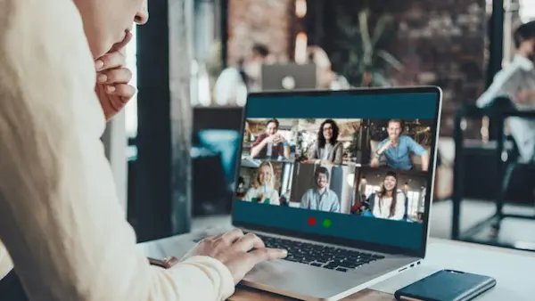 Trabajo híbrido mostrando a una persona escribiendo en una pizarra mientras un equipo se conecta virtualmente en una pantalla.